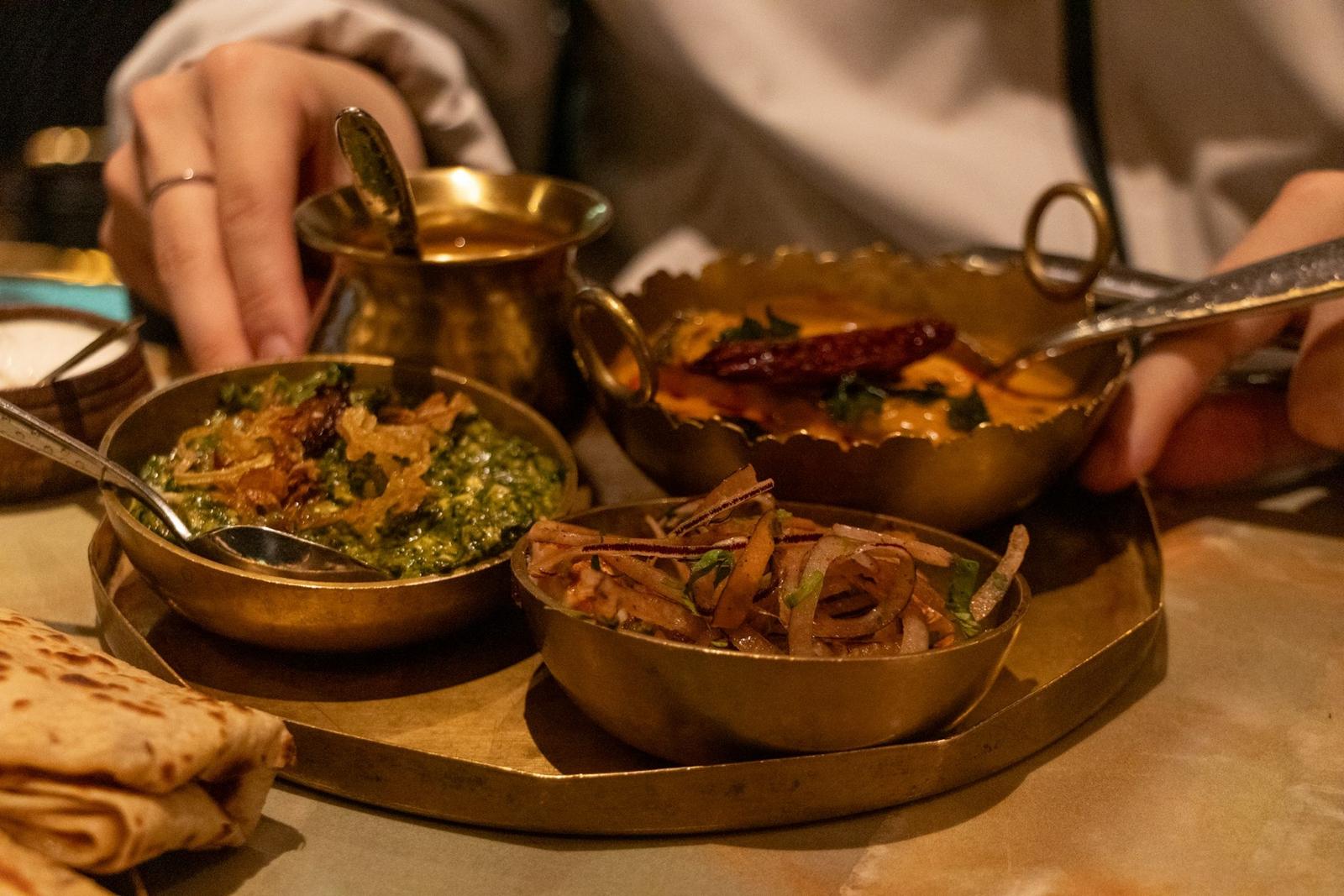 Hands reaching for brass katoris of curry at a tasting table